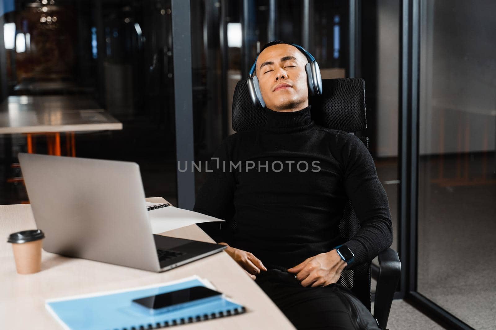 Overtime working. Asian man with closed eyes is resting at work at laptop. Tired male programmer taking a break due to overtime work by Rabizo