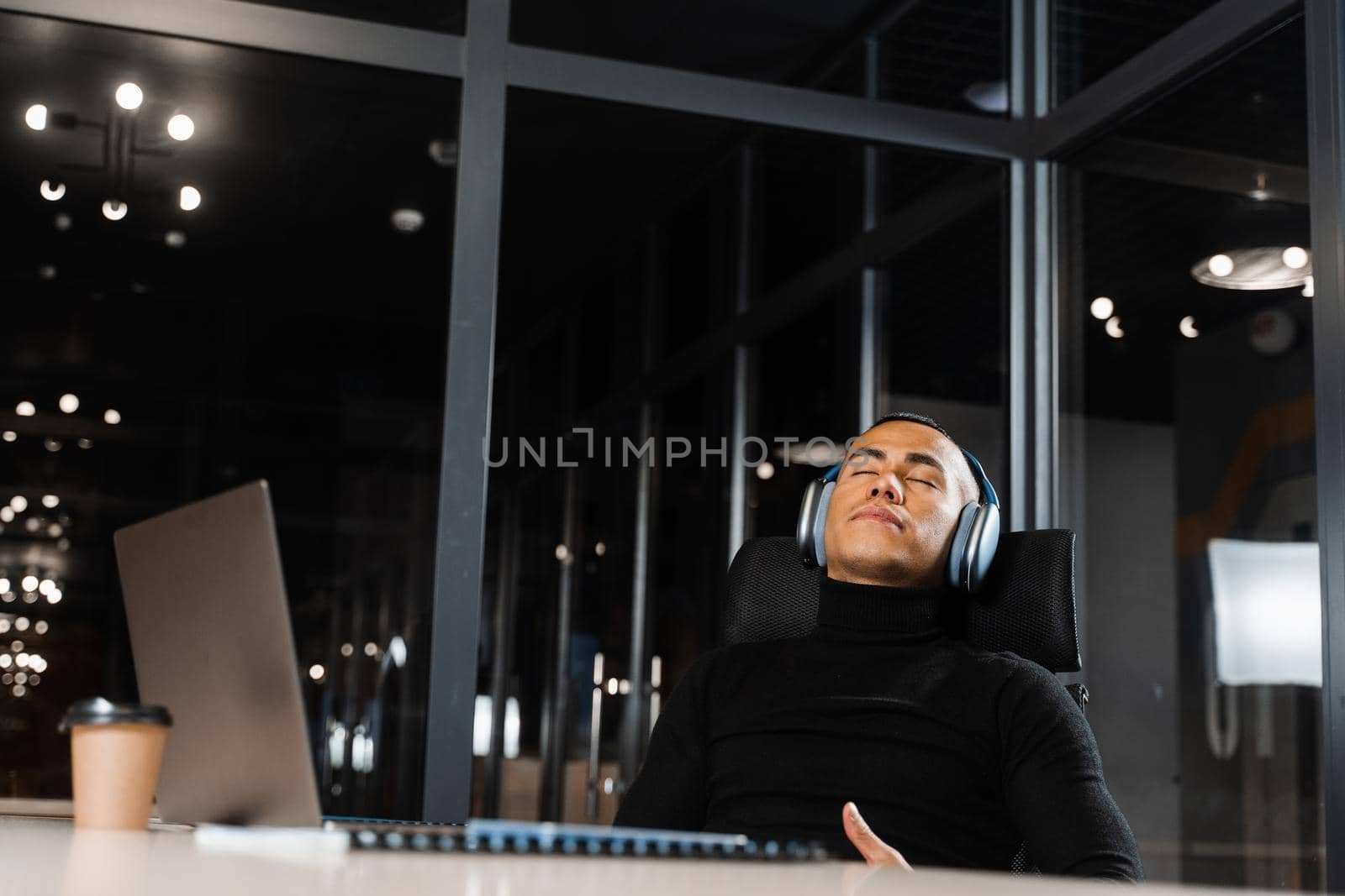 Overtime working. Asian man with closed eyes is resting at work at laptop. Tired male programmer taking a break due to overtime work by Rabizo