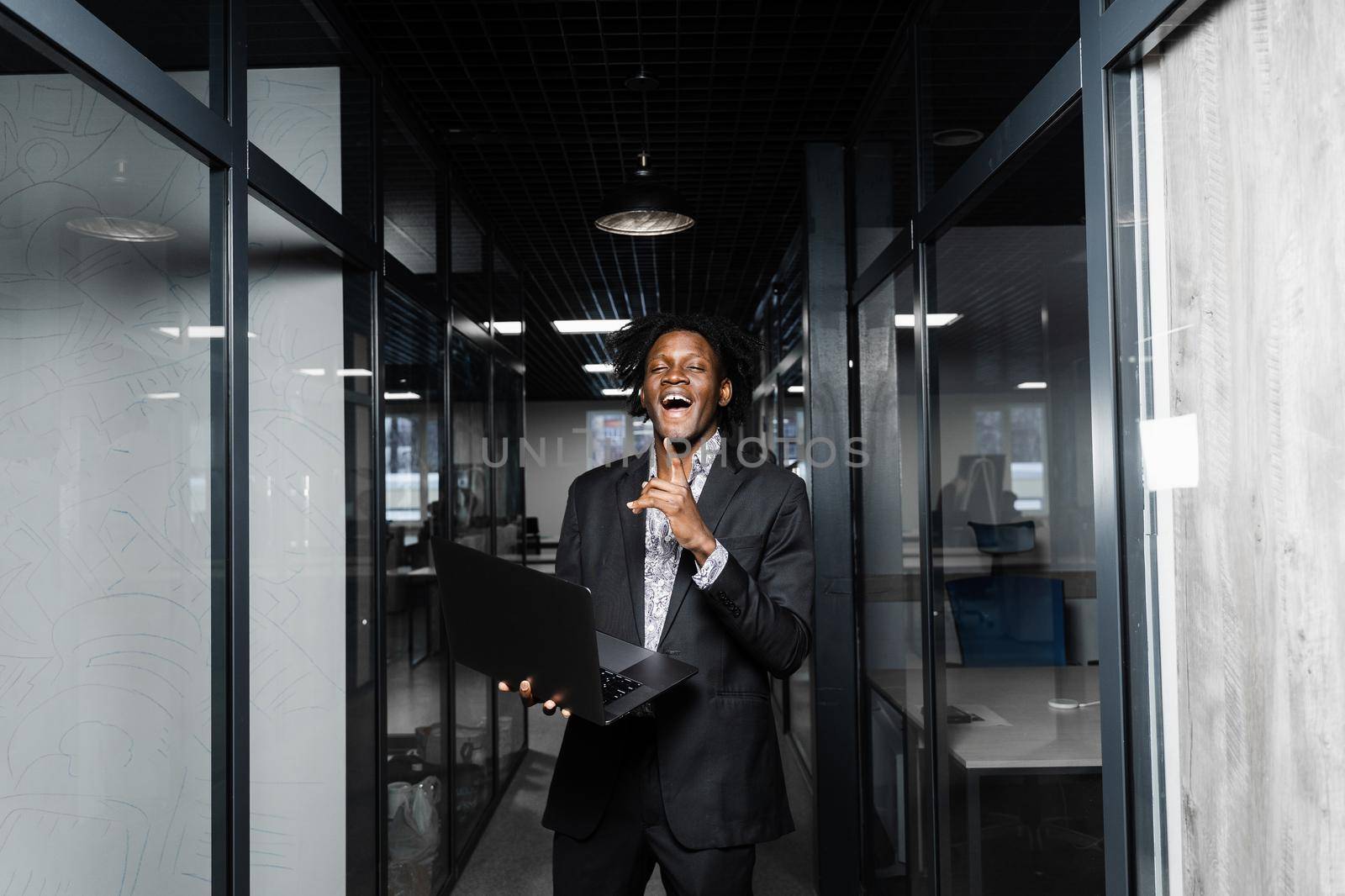 Cheerful black hipster man with laptop is pointing fingers up and smiling in office