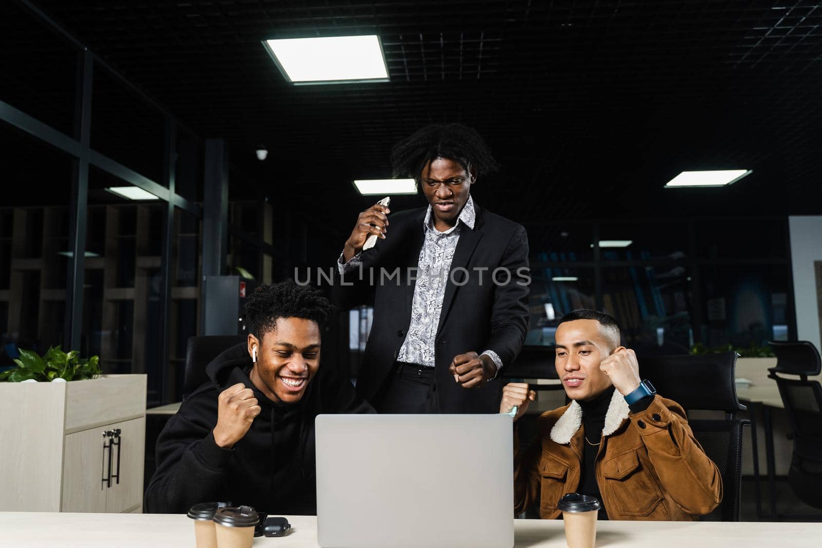 Multiethnic mixed race happy colleagues witl laptop. Black african and asian men raise their hands up, rejoice in victory and shout because they won an online bet. by Rabizo