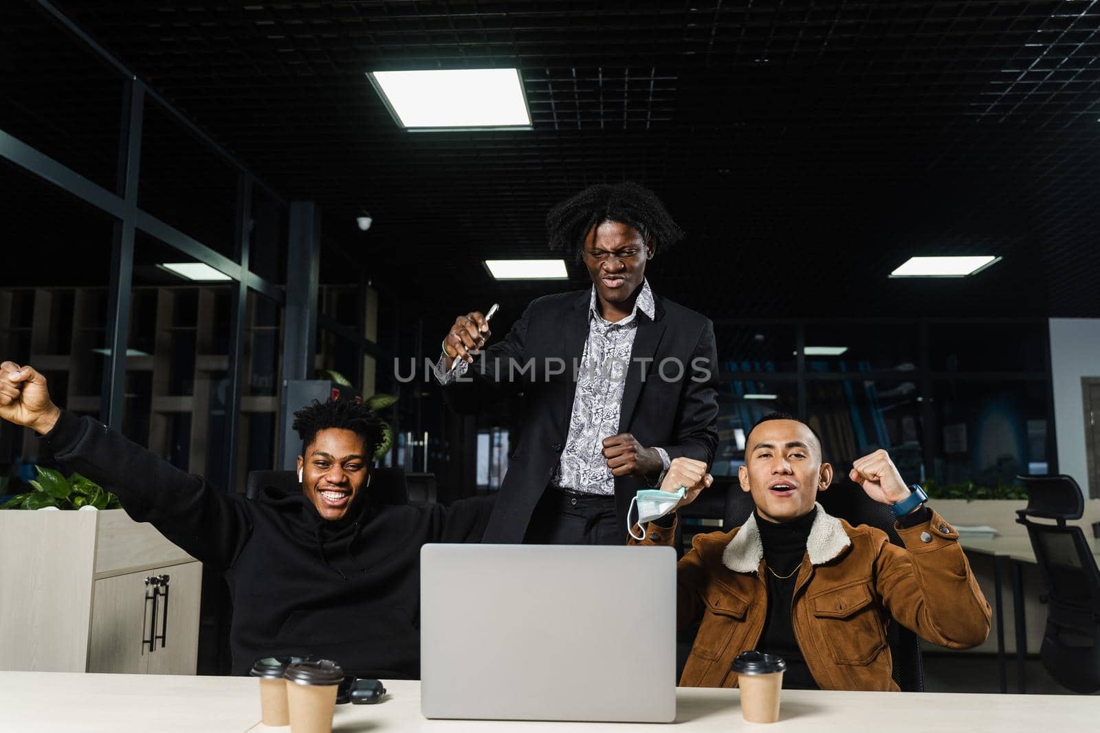 Happy and joyful multiethnic men raise their hands and fists and rejoice in winning in online casino on laptop. Team of black Africans and Asian programmer successful completed project. by Rabizo