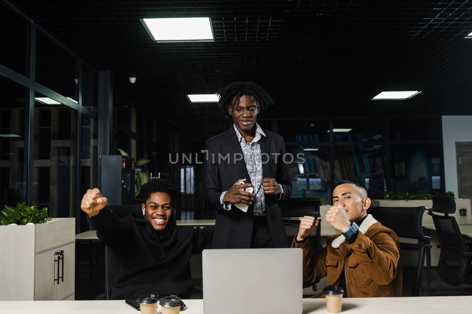 Emotional and joyful black Africans and Asians teamwork together and rejoice at end of online project. Multiethnic colleagues working on laptop, raising their hands, fists and shouting loudly. by Rabizo