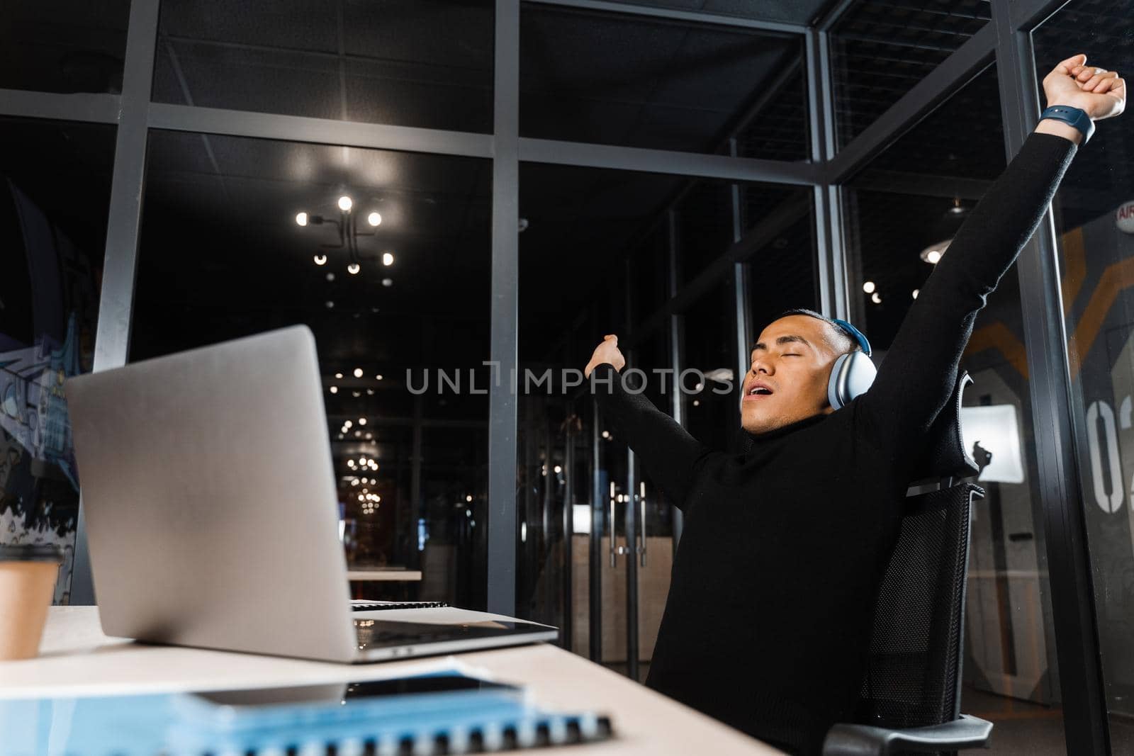 Tired and sleepy Asian man raises his hands up and yawns at the workplace. Work online overtime. Man wants to sleep at work. by Rabizo