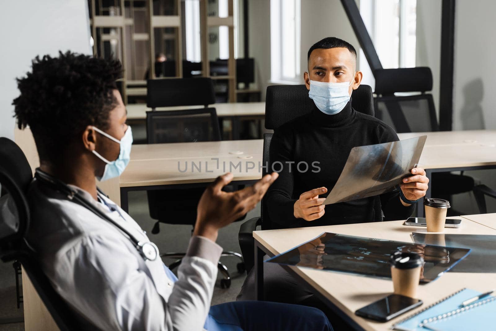 Handsome asian patient with x-ray image of fracture of foot bones at black doctor consultation. African surgeon discussing x-ray with patient