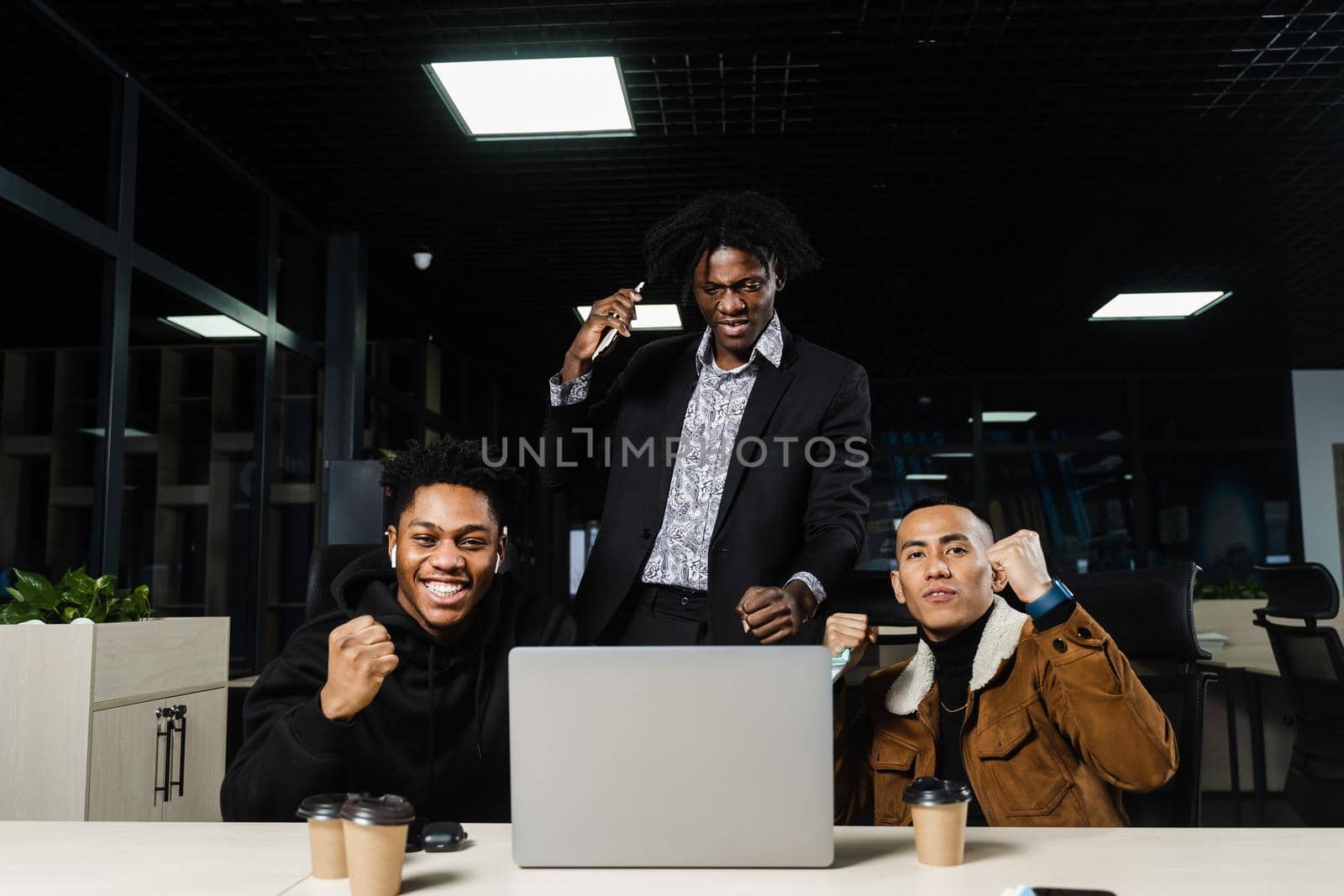 Happy and joyful multiethnic men raise their hands and fists and rejoice in winning in online casino on laptop. Team of black Africans and Asian programmer successful completed project