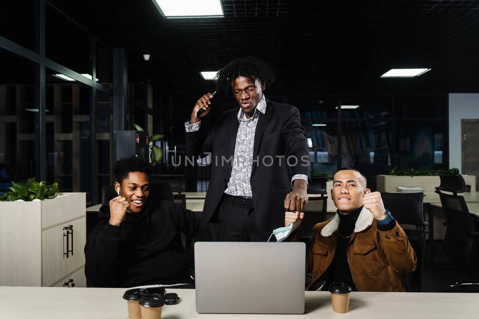 Multiethnic mixed race happy colleagues witl laptop. Black african and asian men raise their hands up, rejoice in victory and shout because they won an online bet