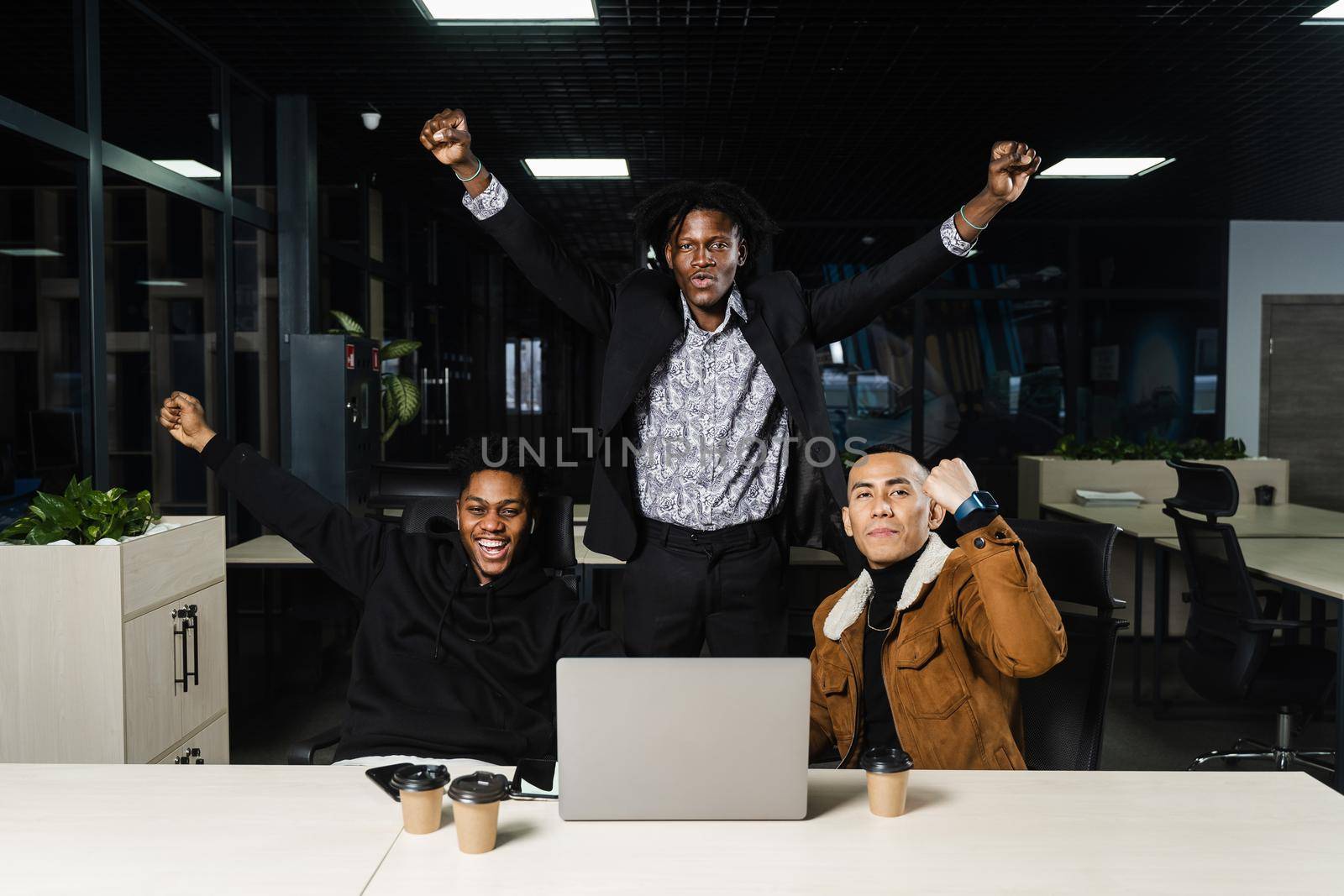 Happy and joyful multiethnic men raise their hands and fists and rejoice in winning in online casino on laptop. Teambuilding of black Africans and Asian programmers