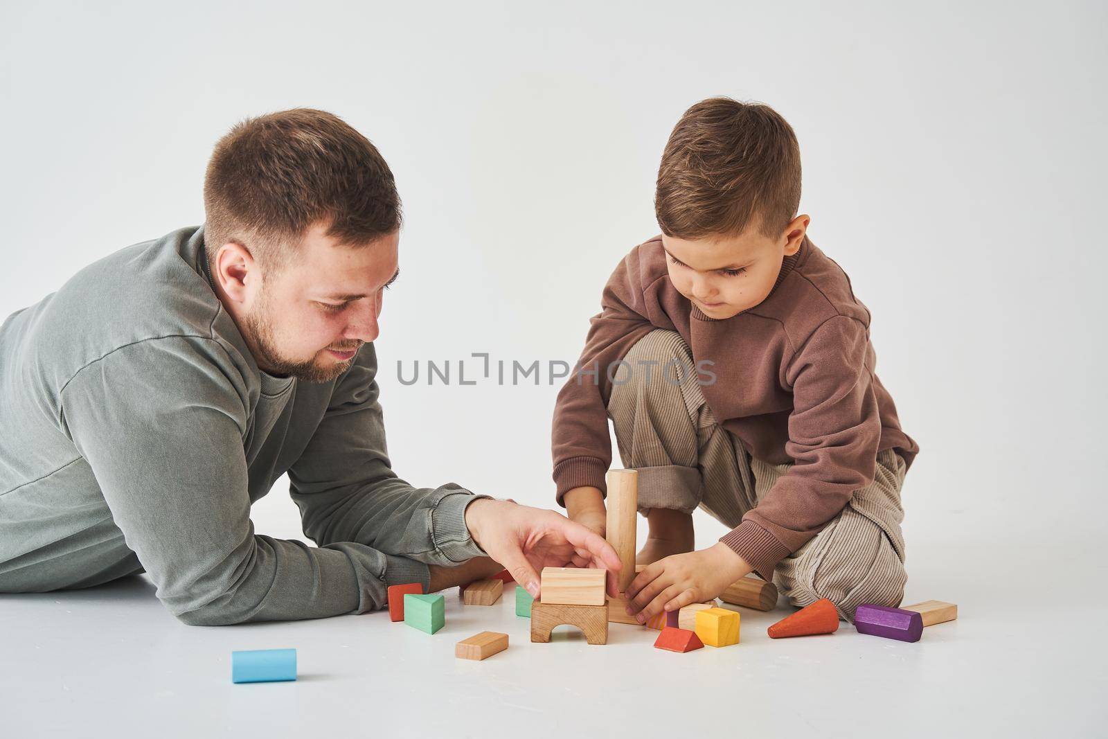 Caring dad plays with cheerful son with toy wooden cubes on white background. Fatherhood and child care. by Rabizo