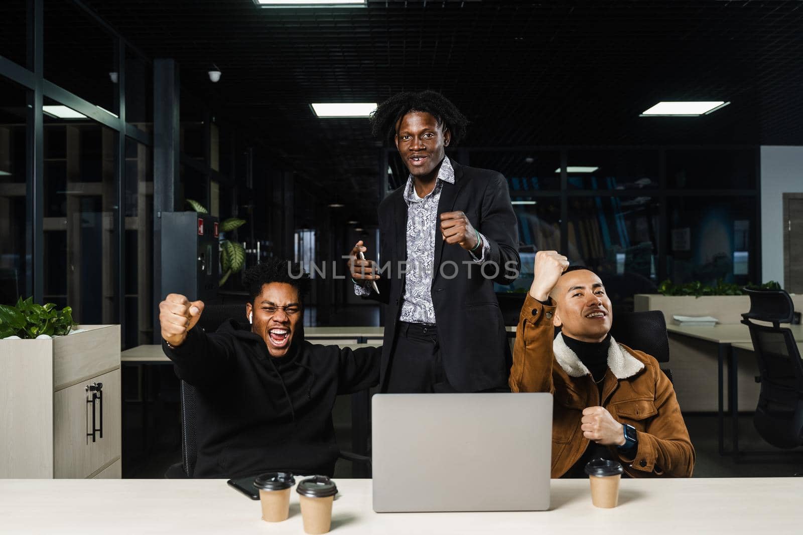 Emotional and joyful black Africans and Asians teamwork together and rejoice at end of online project. Multiethnic colleagues working on laptop, raising their hands, fists and shouting loudly. by Rabizo