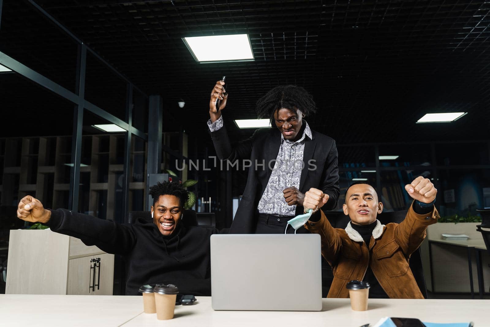 Happy and joyful multiethnic men raise their hands and fists and rejoice in winning in online casino on laptop. Team of black Africans and Asian programmer successful completed project. by Rabizo