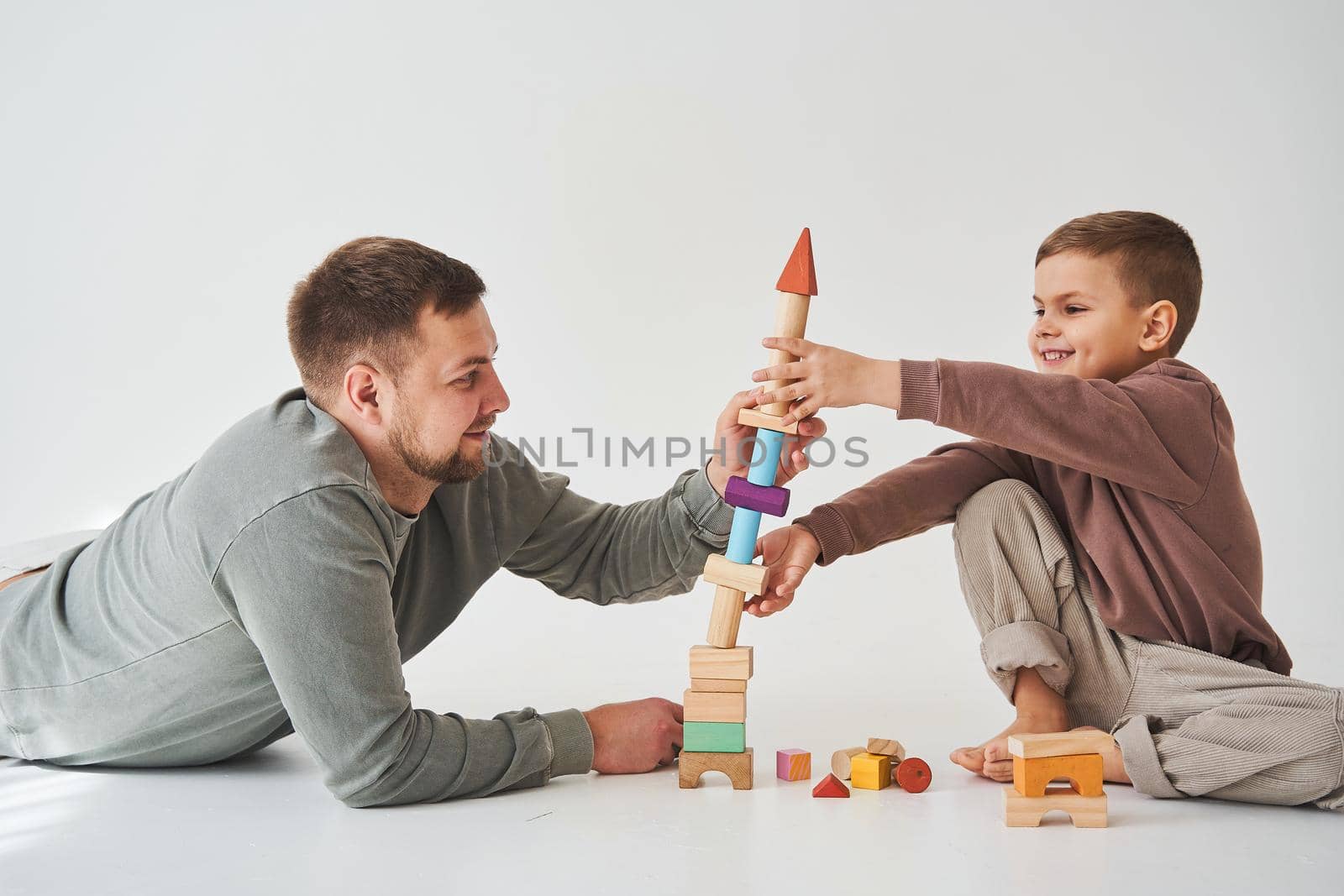 Caring dad helps his son to play on the floor on white background. Father and child build tower of colorful wooden bricks and have fun together. by Rabizo