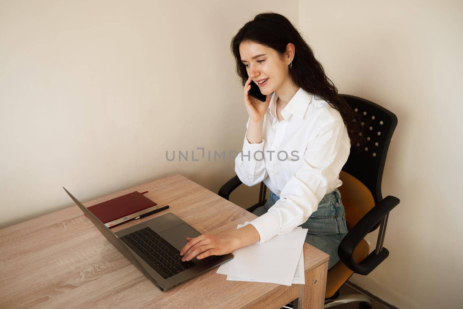 Attractive girl with laptop and papers working online and write notes on paper and talking by phone. Confident business woman work at home. by Rabizo