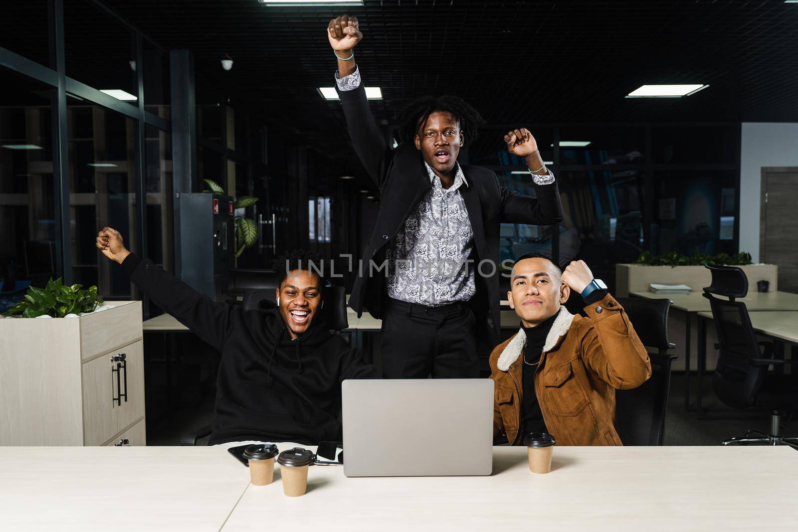 Happy and joyful multiethnic men raise their hands and fists and rejoice in winning in online casino on laptop. Teambuilding of black Africans and Asian programmers