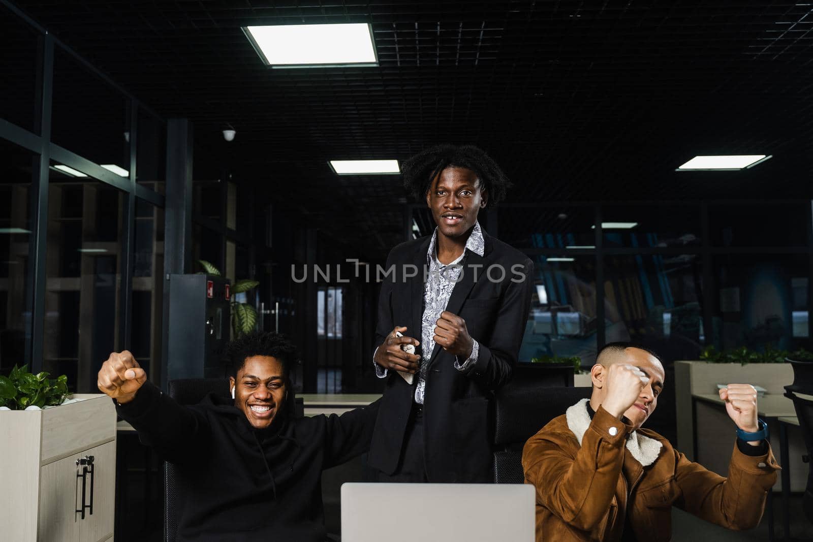 Emotional and joyful black Africans and Asians teamwork together and rejoice at end of online project. Multiethnic colleagues working on laptop, raising their hands, fists and shouting loudly