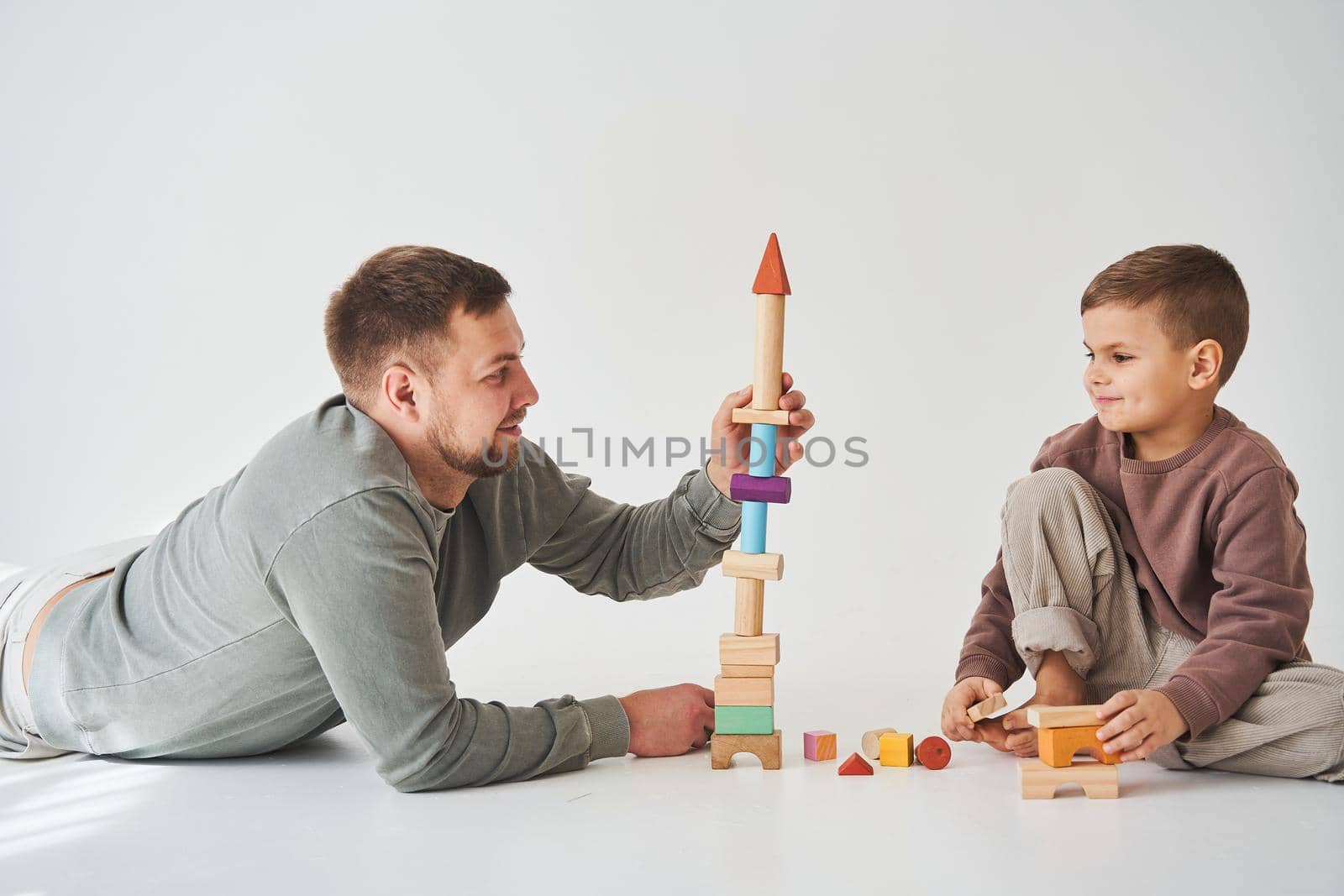 Caring dad helps his son to play on the floor on white background. Father and child build tower of colorful wooden bricks and have fun together. by Rabizo