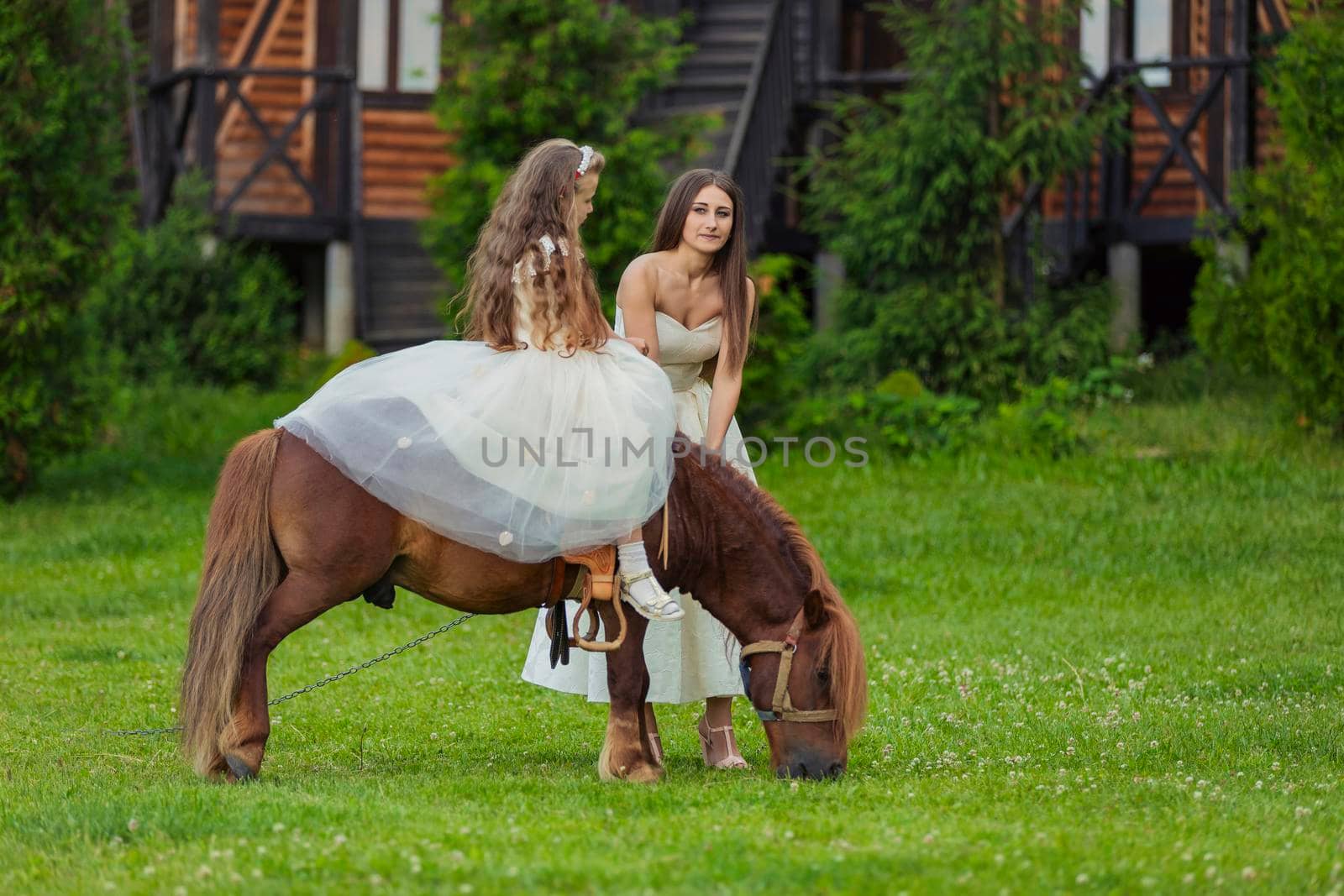 mother rides her daughter on a pony on a green lawn