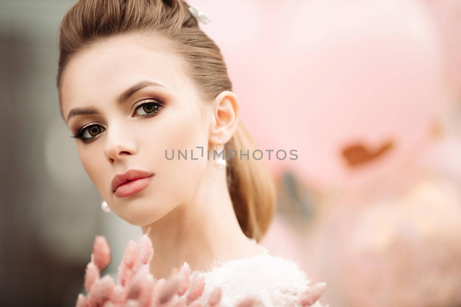 Headshot of gorgeous brunette woman with flawless skin, high hairdo, professional make up and bunch of pink flowers looking at camera. Unfocused pink background.