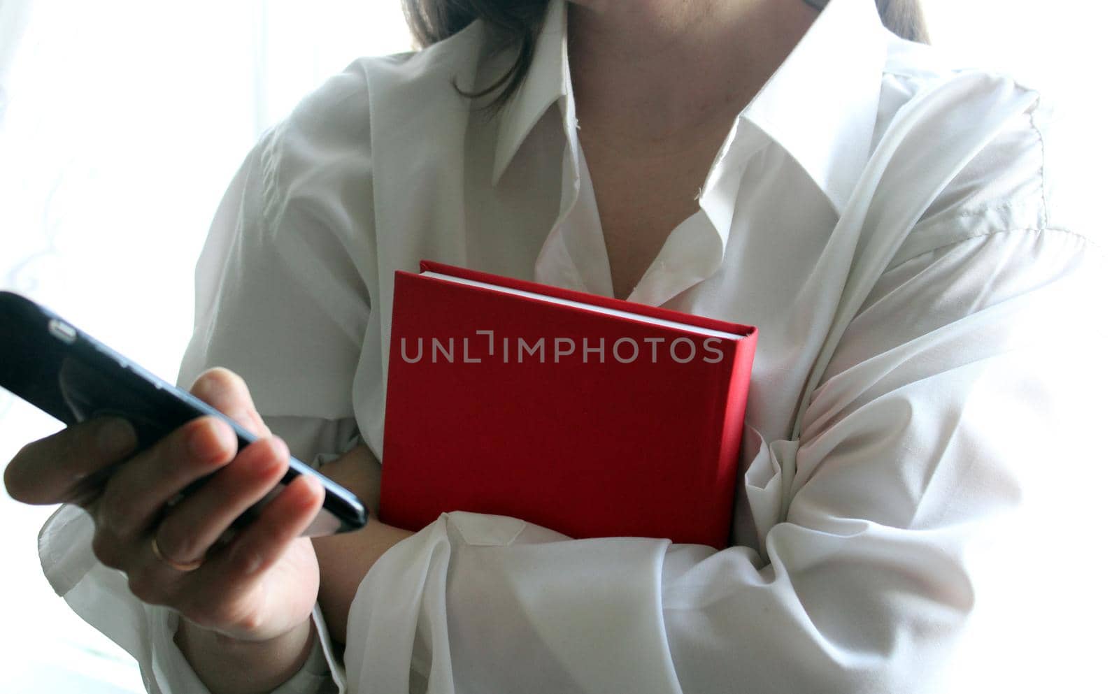 A beautiful girl in a white shirt holds a smartphone and a red book in her hands..