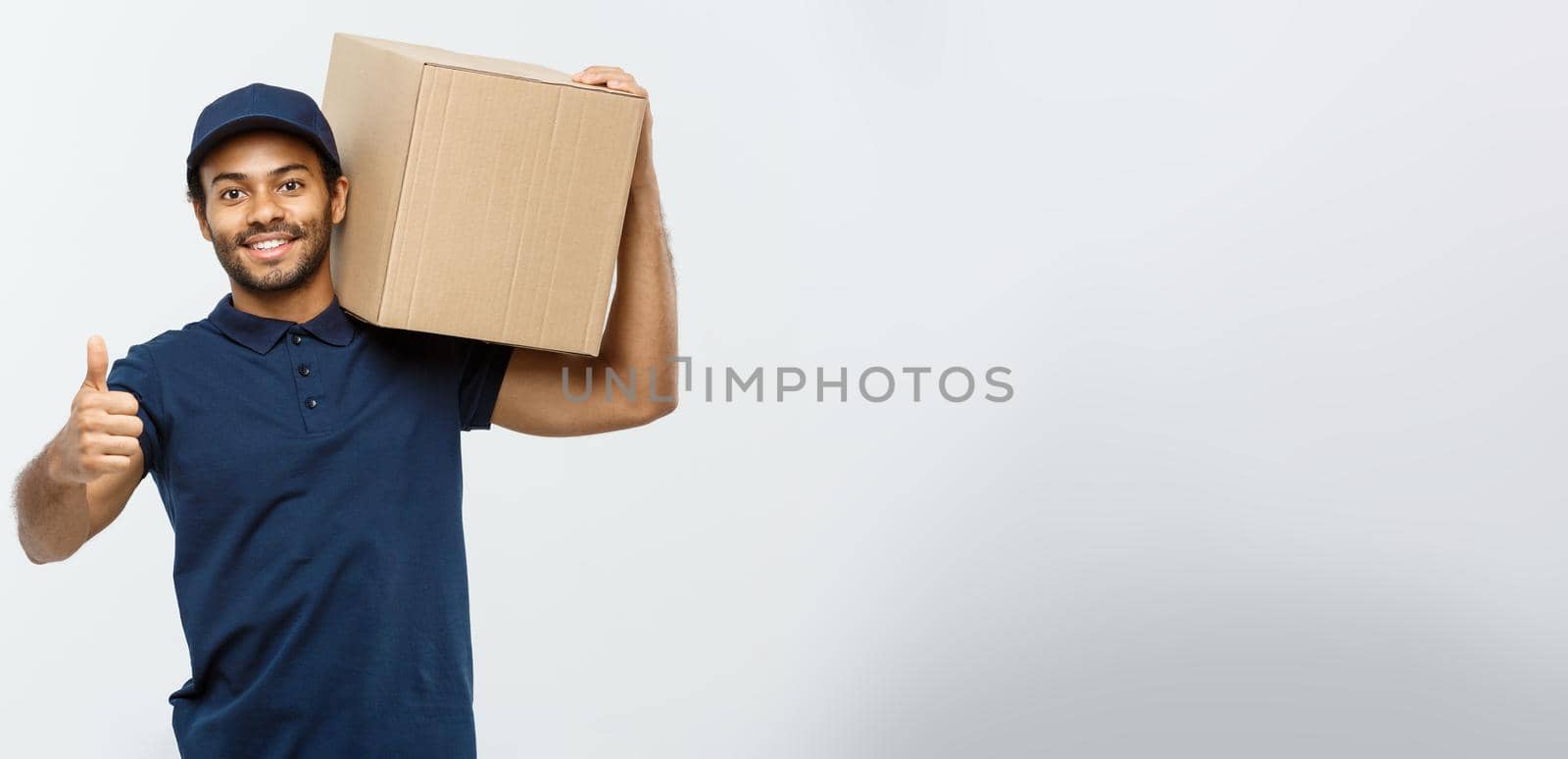 Delivery Concept - Portrait of Happy African American delivery man holding a box package and showing thumps up. Isolated on Grey studio Background. Copy Space