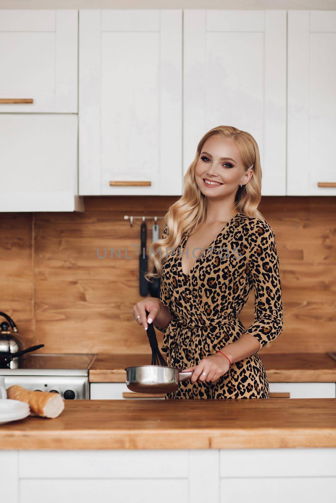 Stock photo portrait of stunning elegant blonde woman with hairstyle wearing leopard printed dress cooking in the kitchen at home. She looks gorgeous and preparing breakfast in the kitchen.