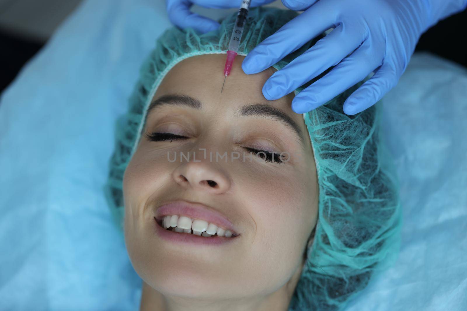 Portrait of smiling lady gets injection of botox in her forehead, cosmetologist using syringe with liquid. Cosmetology, beauty treatment, wellness concept