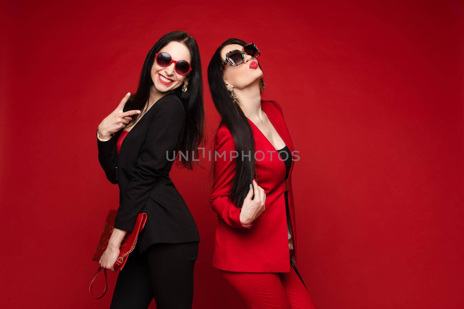 Two young fashion stylish female model in sunglasses smiling posing isolated at red studio by StudioLucky