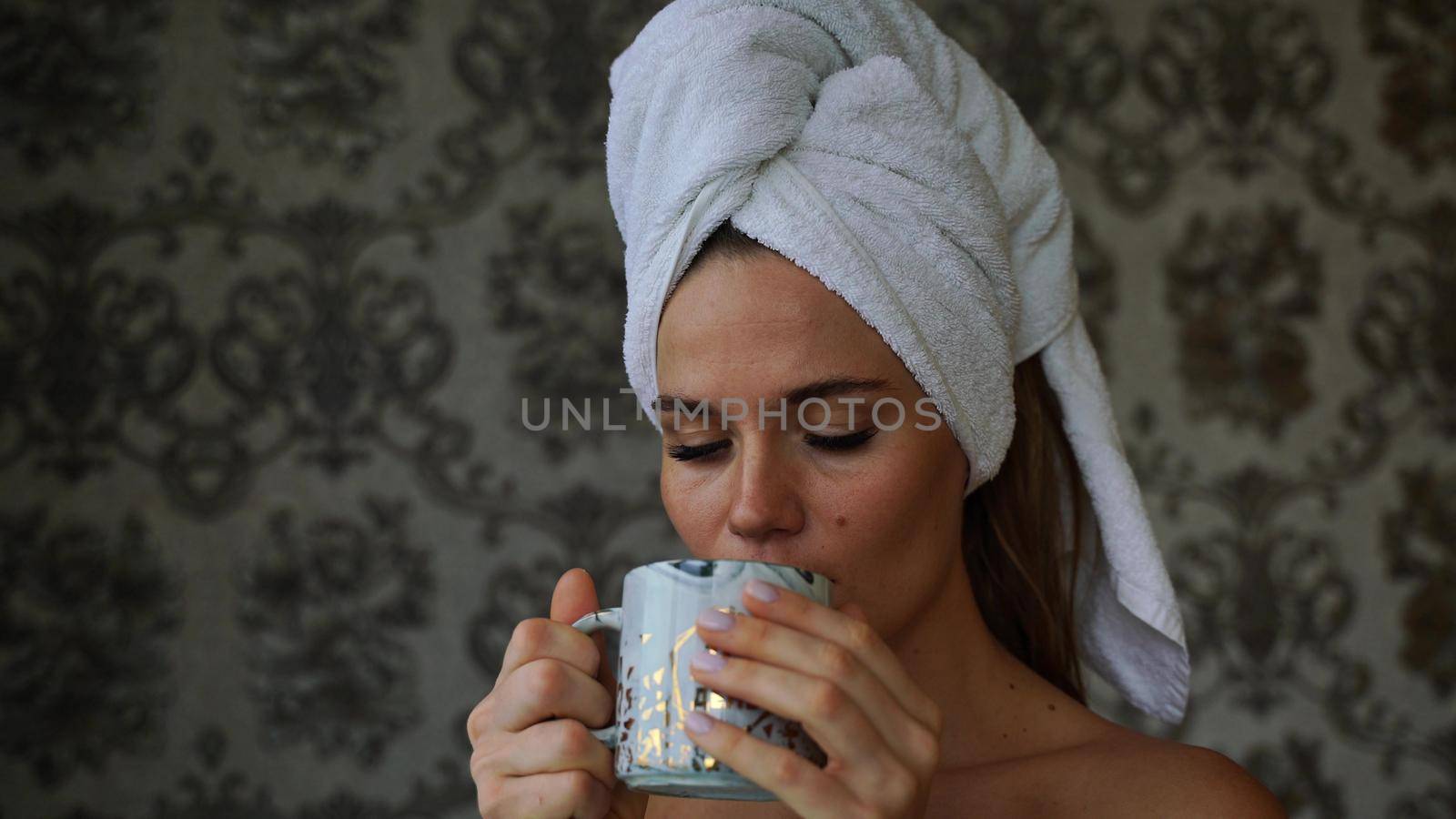 Middle-aged woman looks good with bare shoulders in a white towel on her head holds a cup and drinks coffee or tea against the wall.