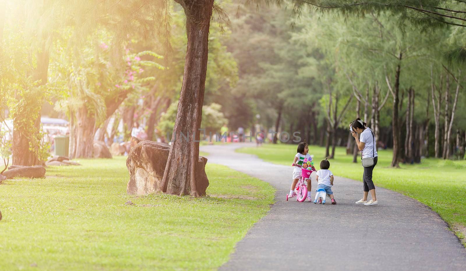 Family in park concept. Mother and her daughter play at park together. Mother shot her daugther with camera. Little girls relax with little bicycle on road in park.
Happy young mother shot picture her daugther.