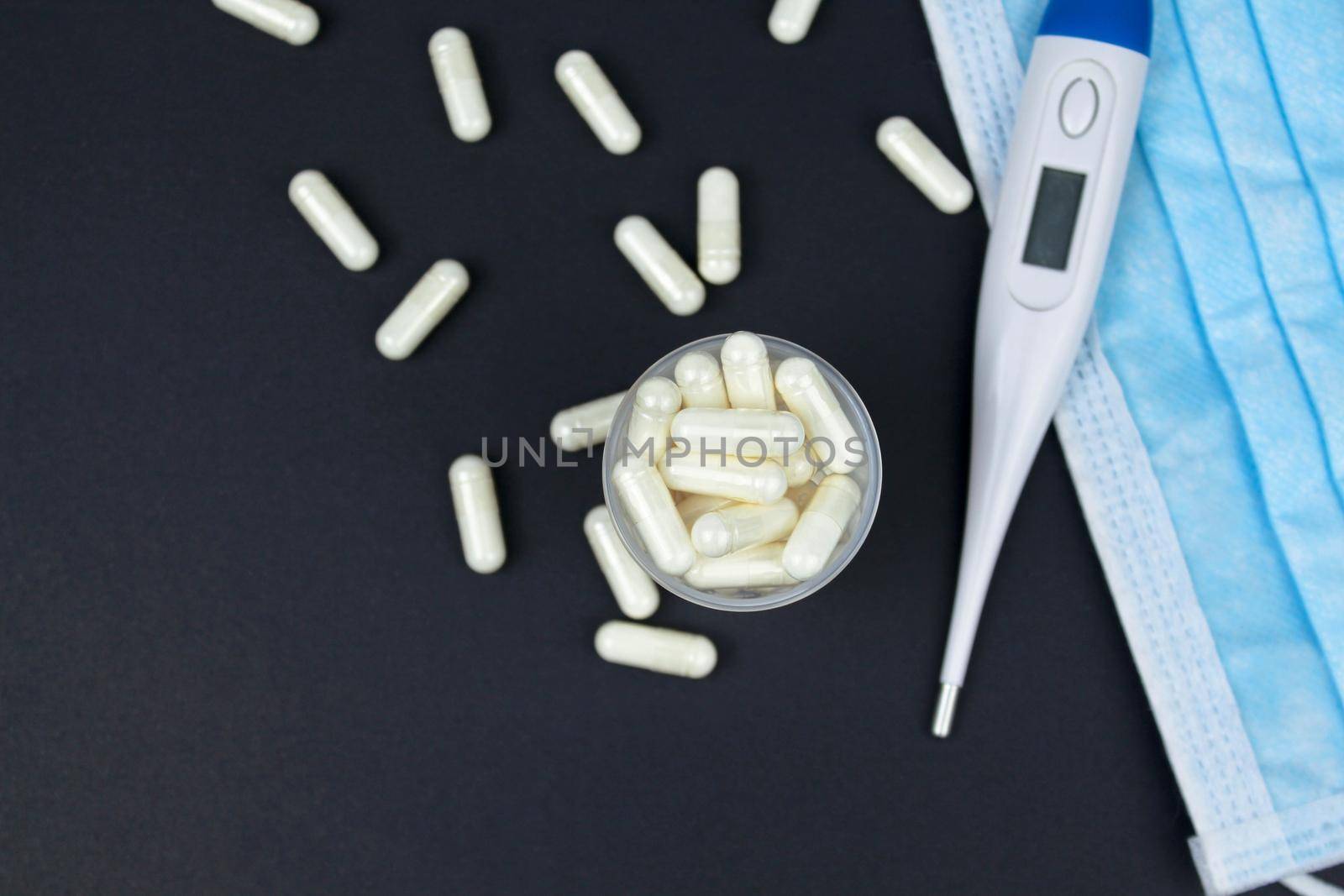 White electronic thermometer with packs of tablets and blue medical face mask on black background. Selective focus.