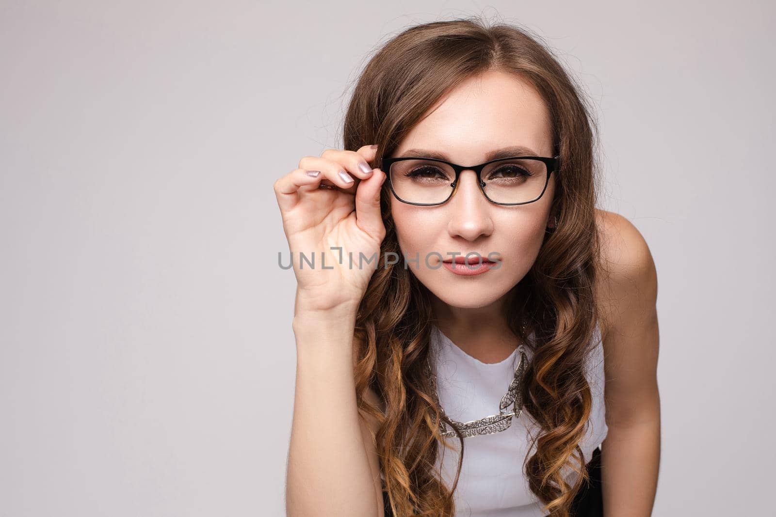 Front view of shocked long haired woman keeping glasses and looking with big eyes in camera. Young female in white shirt standing and posing on grey isolated background. Concept of surprise.