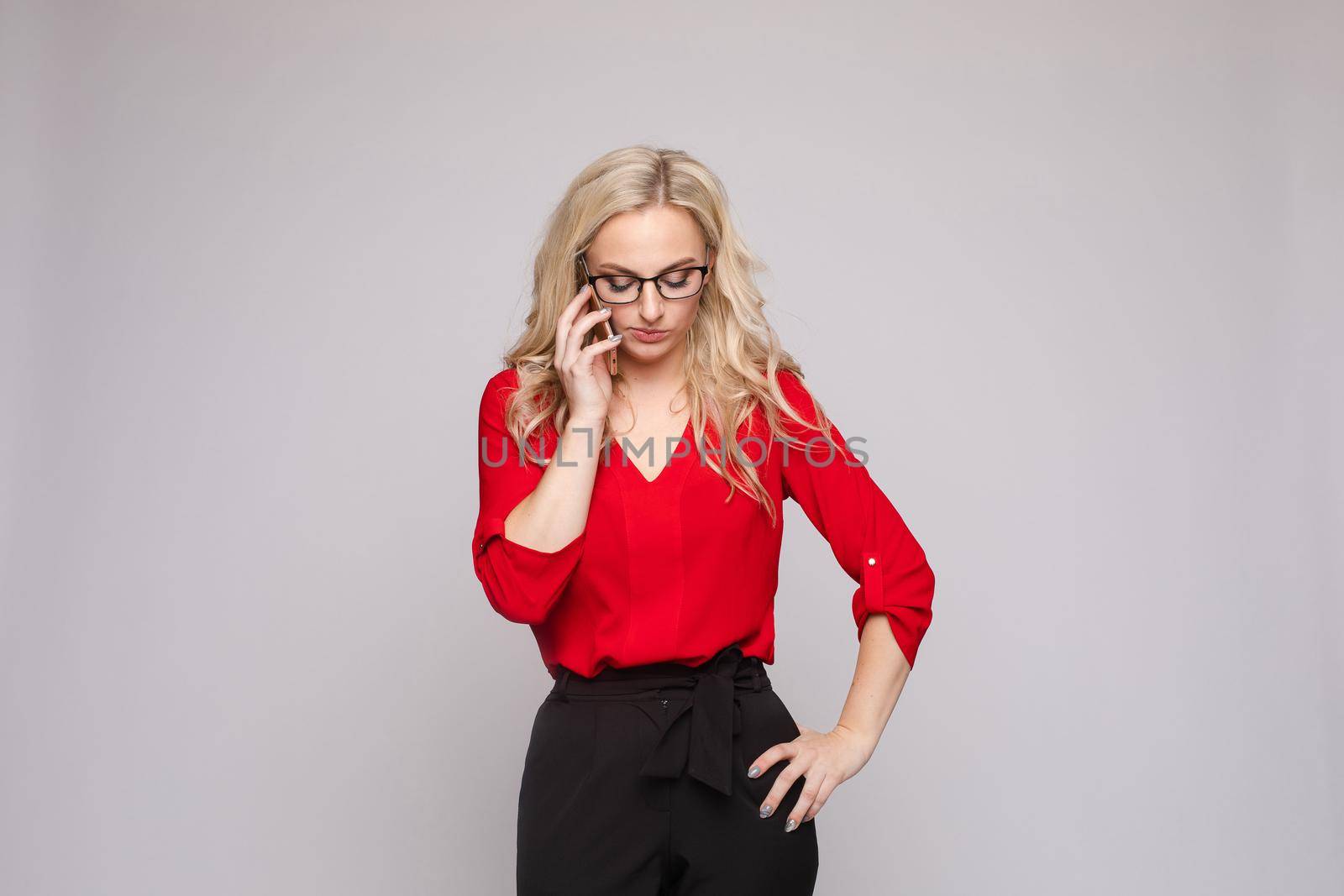 Front view of smiling woman wearing red blouse and skirt keeping phone and laughing on isolated background. Cheerful blonde messaging and communicating in studio. Concept of technology and business.