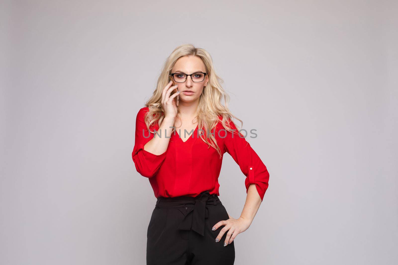 Front view of smiling woman wearing red blouse and skirt keeping phone on isolated background. Cheerful blonde messaging and communicating in studio. Concept of technology and business.