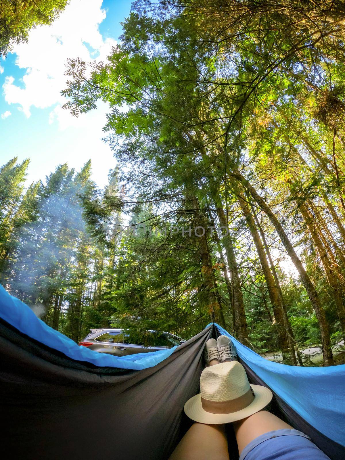 Woman relaxing in the hammock by the lake in the forest, POV view of legs in trekking boots with straw hat. Wanderlust concept scene. by JuliaDorian