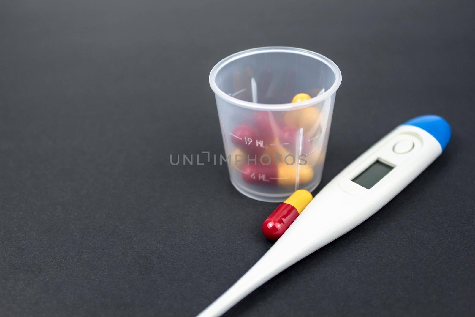 Medical capsules, tablets and clinical thermometer inside measuring cup on black background. Side view