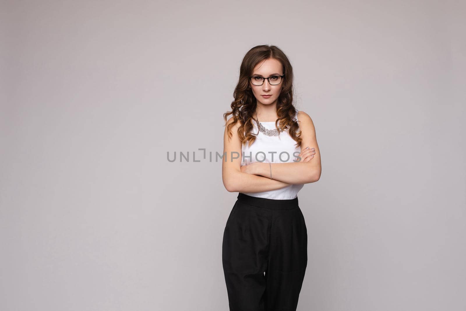 Front view of shocked long haired woman keeping glasses and looking with big eyes in camera. Young female in white shirt standing and posing on grey isolated background. Concept of surprise.
