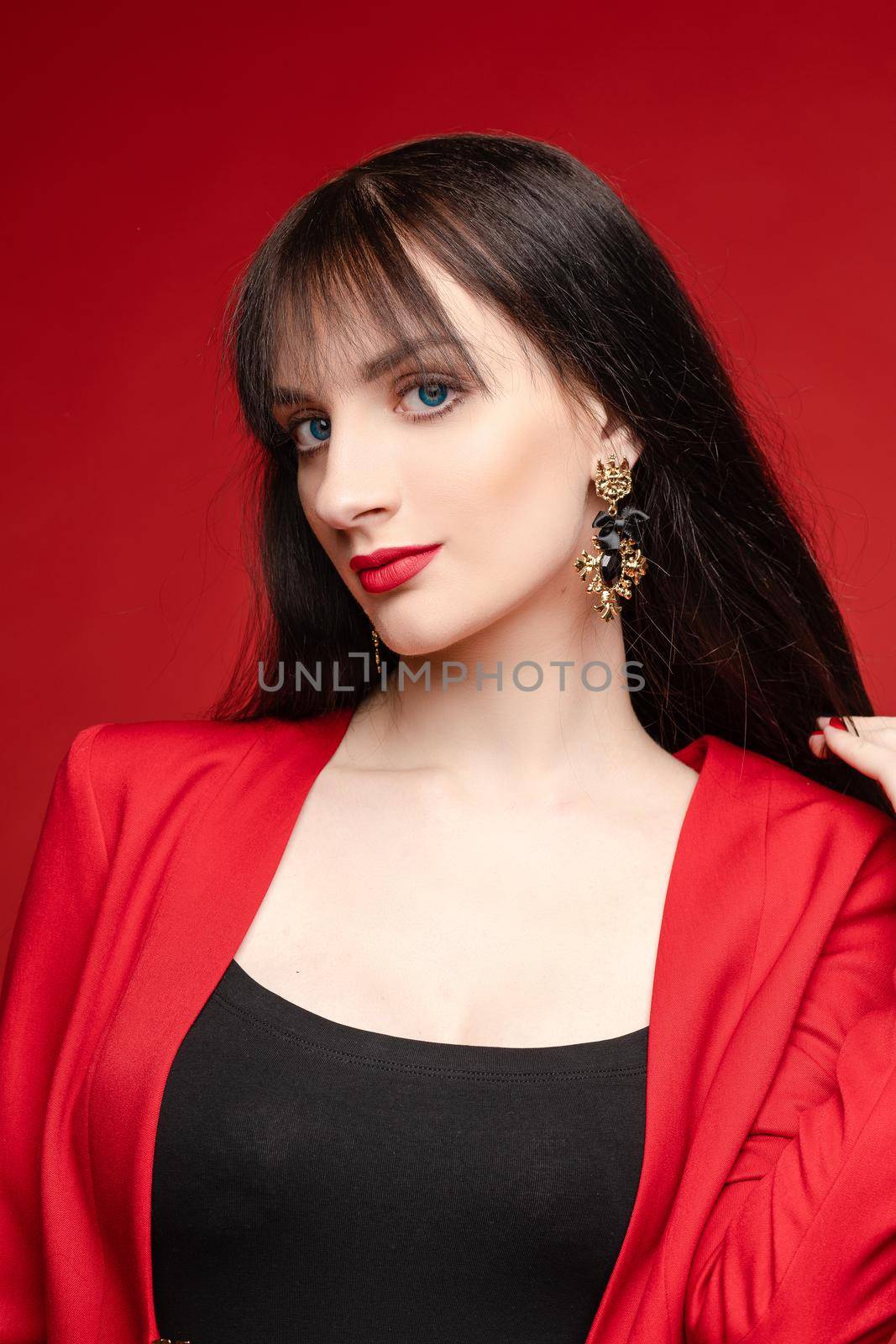 Side view of glamorous brunette posing in red smart suit and heels on red isolated background in studio. Young woman keeping glasses in hand and looking down. Concept of beauty and fashion.