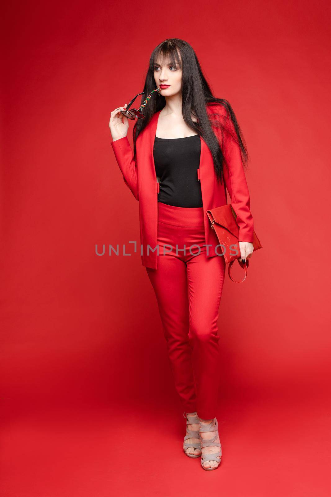 Side view of glamorous brunette posing in red smart suit and heels on red isolated background in studio. Young woman keeping glasses in hand and looking down. Concept of beauty and fashion.