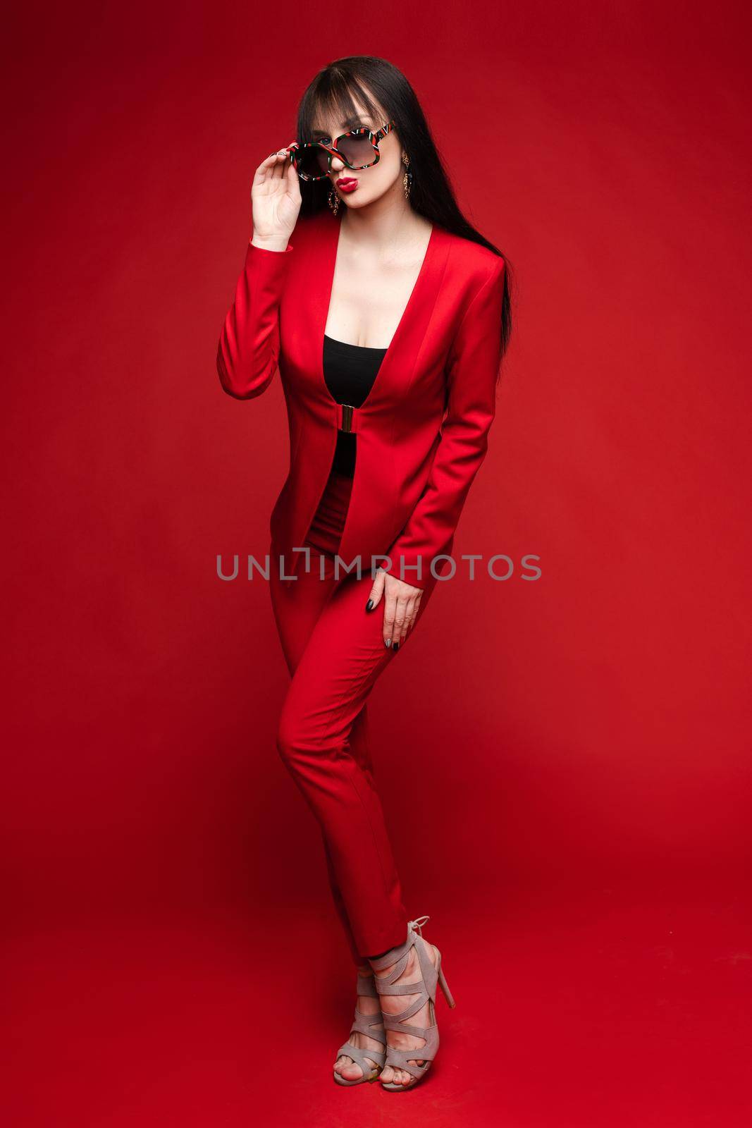 Side view of glamorous brunette posing in red smart suit and heels on red isolated background in studio. Young woman keeping glasses in hand and looking down. Concept of beauty and fashion.