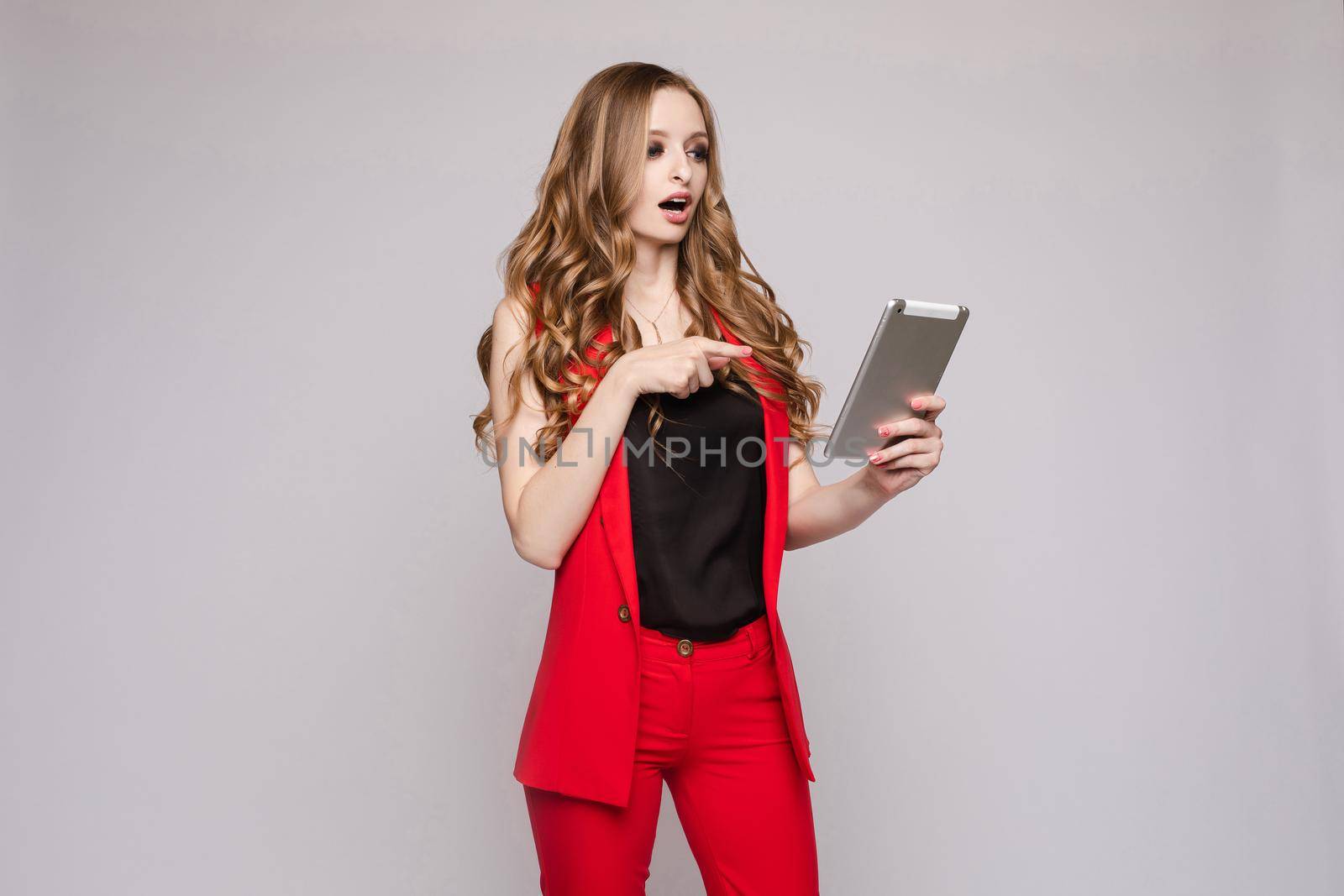 Young beautiful girl holding silver tablet and reading book on it. Brunette lady in red jacket and pants posing with electronic device. Elegant business woman in costume seriously checking email.