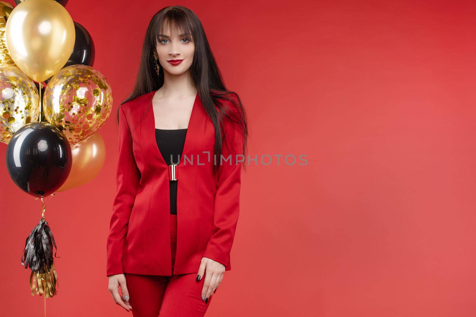 Young brunette model with blue eyes posing in fashionable clothes. Beautiful girl with long hair in elegant red costume looking at camera. Gorgeous lady standing near bunch of black and gold balloons.