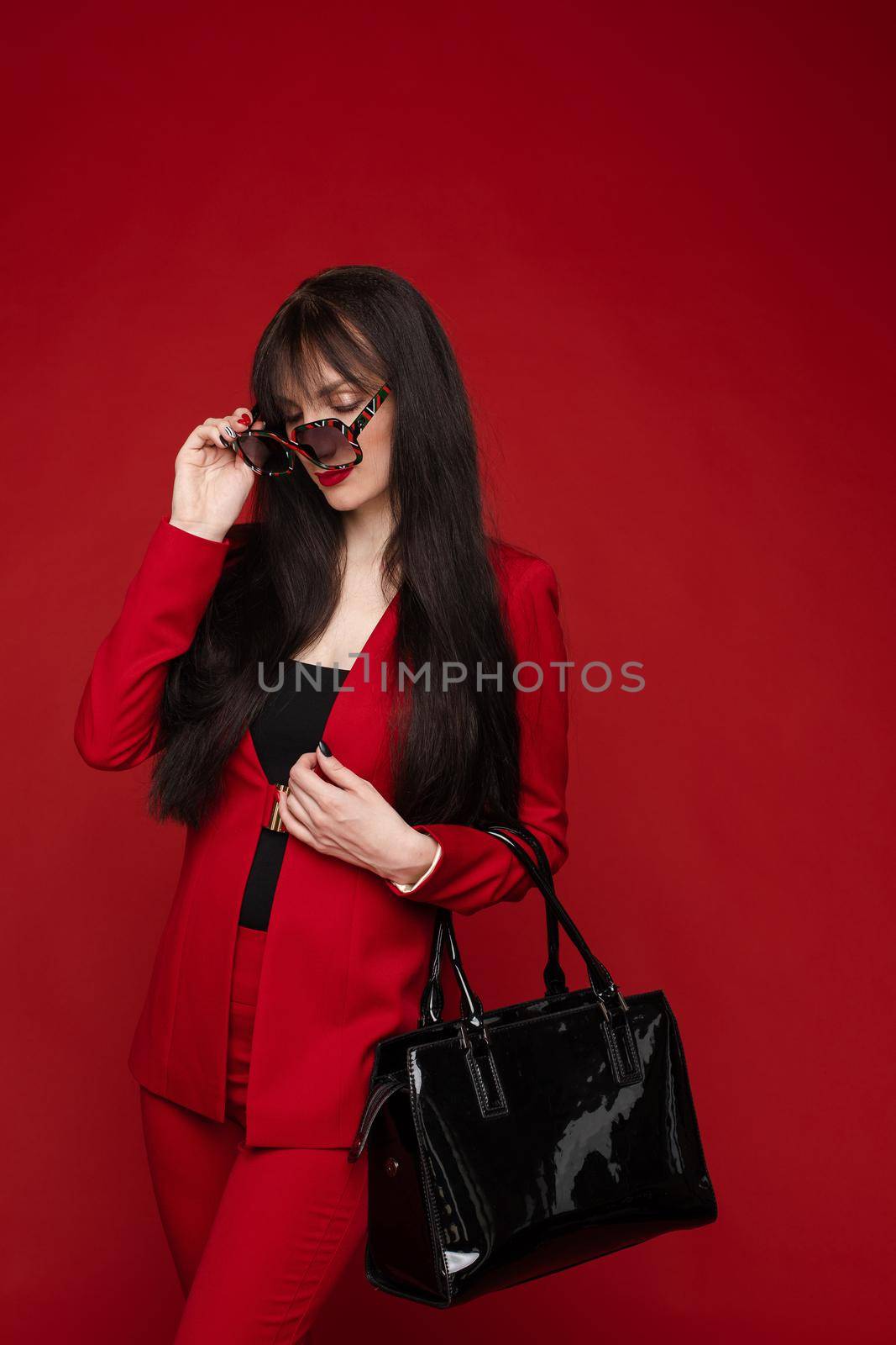 Portrait of young beautiful woman thoughtfully looking down. Brunette model in elegant costume posing at camera . Pretty lady in red sunglasses leaning one hand on other and holding it near face.