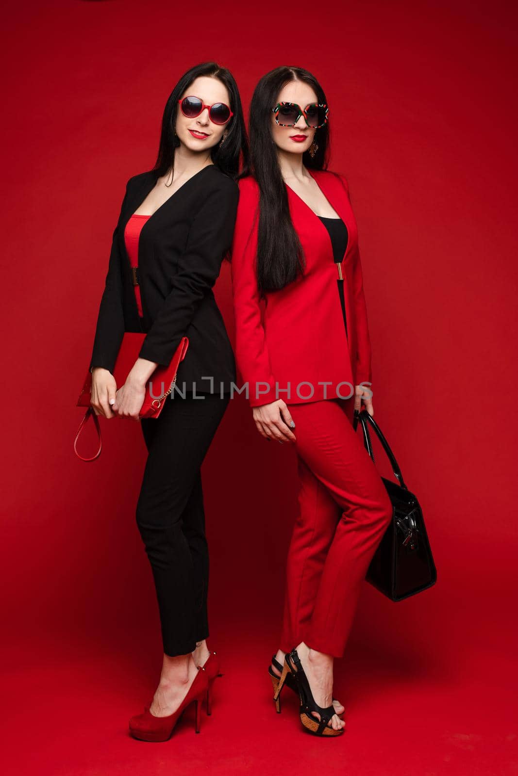 Two young fashion stylish female model in sunglasses smiling posing isolated at red studio by StudioLucky