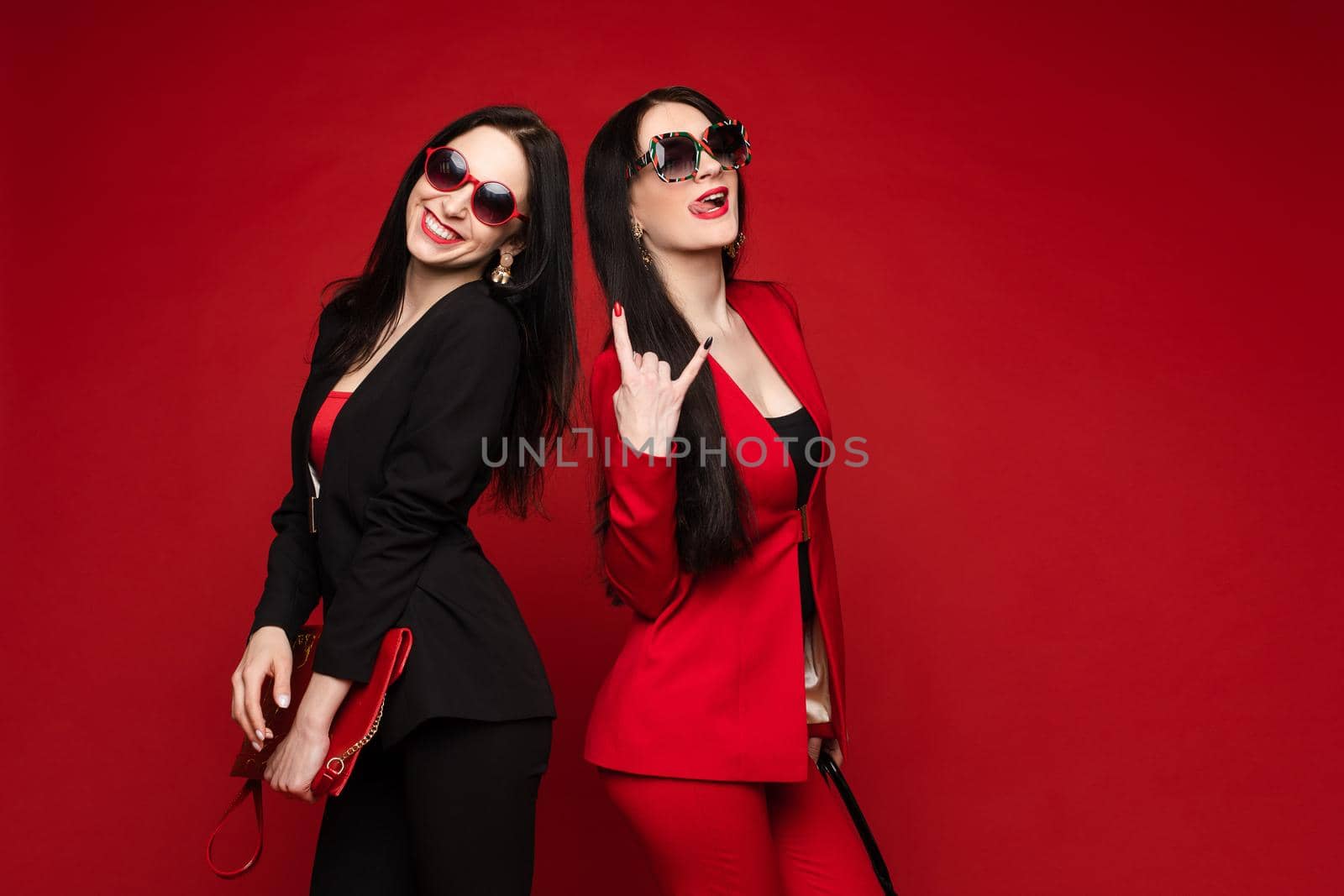 Two young fashion stylish female model in sunglasses smiling posing isolated at red studio background. Adorable happy woman friend relaxing having fun together medium long shot