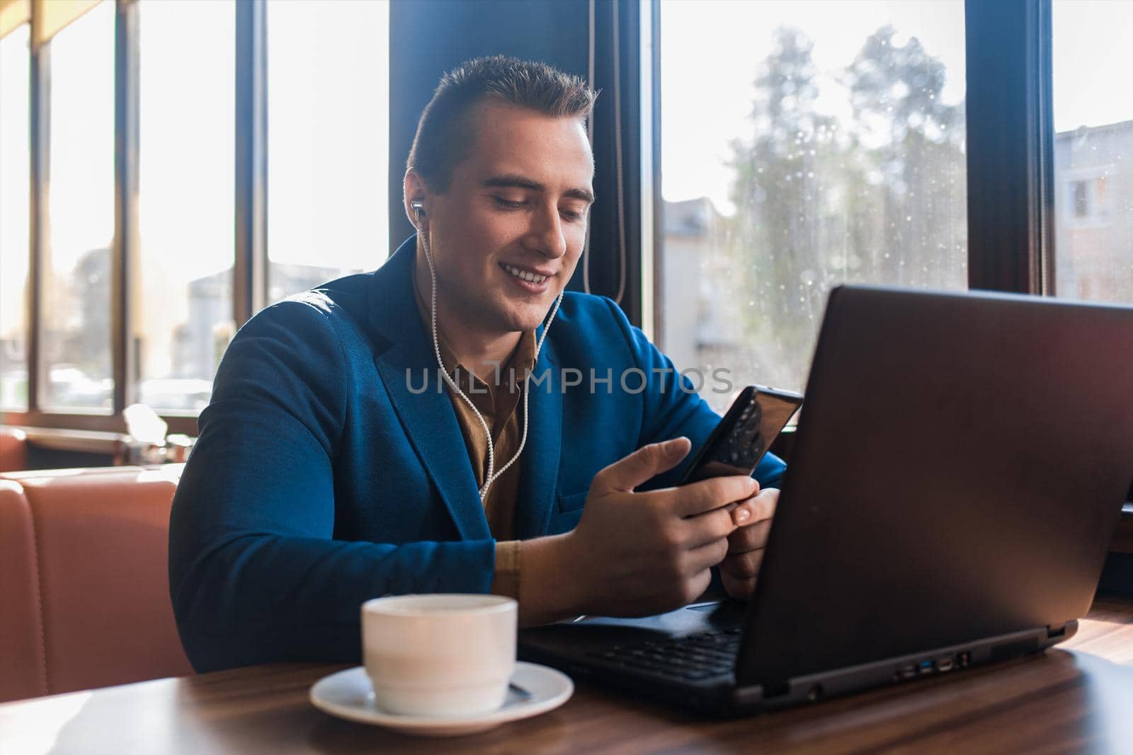 A business happy man stylish smiling businessman in an attractive European-looking suit works in a laptop, listens to music and drinks coffee sitting at a table in a cafe by the window.