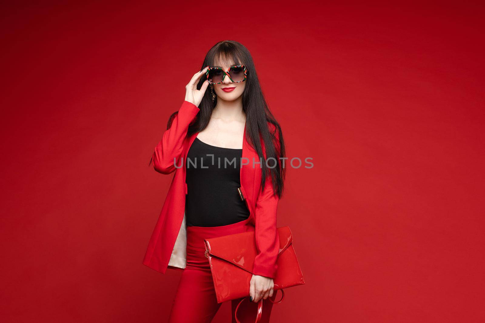 Side view of glamorous brunette posing in red smart suit and heels on red isolated background in studio. Young woman keeping glasses in hand and looking down. Concept of beauty and fashion.
