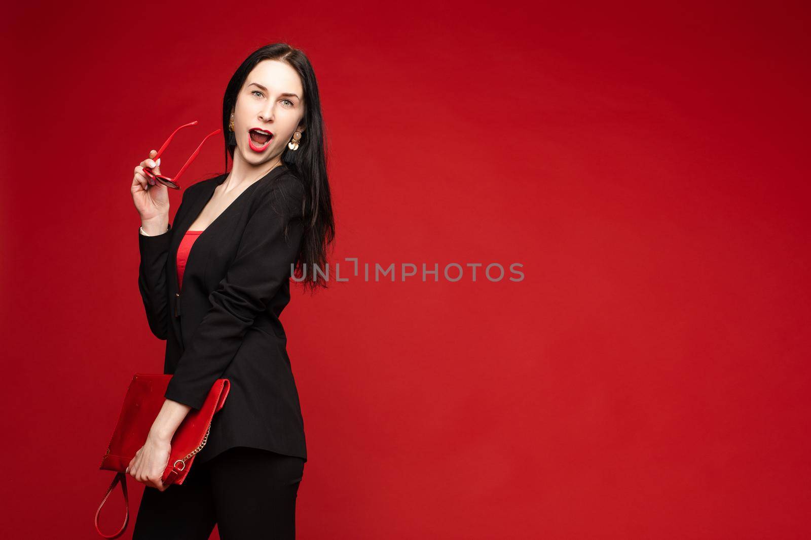 Portrait of young beautiful woman thoughtfully looking down. Brunette model in elegant costume posing at camera . Pretty lady in red sunglasses leaning one hand on other and holding it near face.