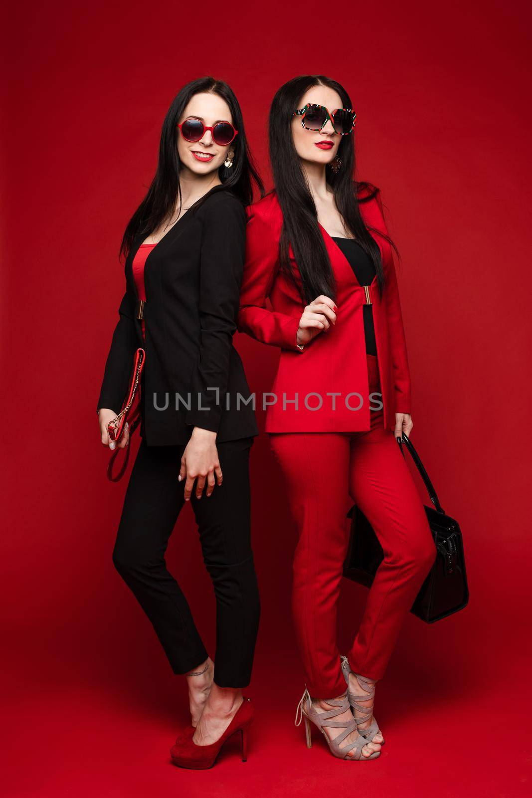 Side view of two brunettes wearing black and red smart suits and glasses standing back to back on isolated background. Stylish women posing and looking at camera in studio. Concept of glamour.