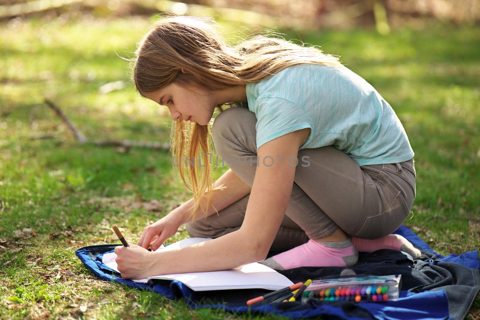 A young adolescent girl writes or colors in a notebook or journal on a blanket on the grass by markvandam