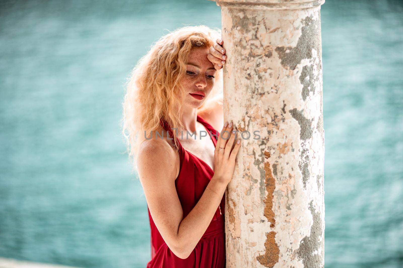 Outdoor portrait of a young beautiful natural redhead girl with freckles, long curly hair, in a red dress, posing against the background of the sea