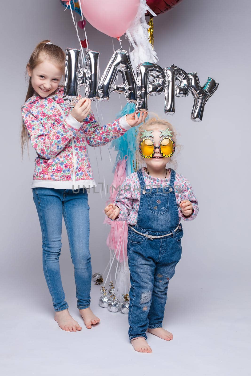 Front view of happy family looking at camera and posing while celebrating birthday party. Young mother keeping little daughter while girl standing near and keeping colorful balloons. Concept of fun.